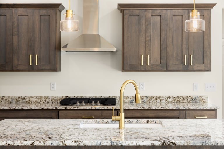 A kitchen detail with a gold faucet on a granite countertop, dark wood cabinets, stainless steel appliances, and gold pendant lights hanging above the island