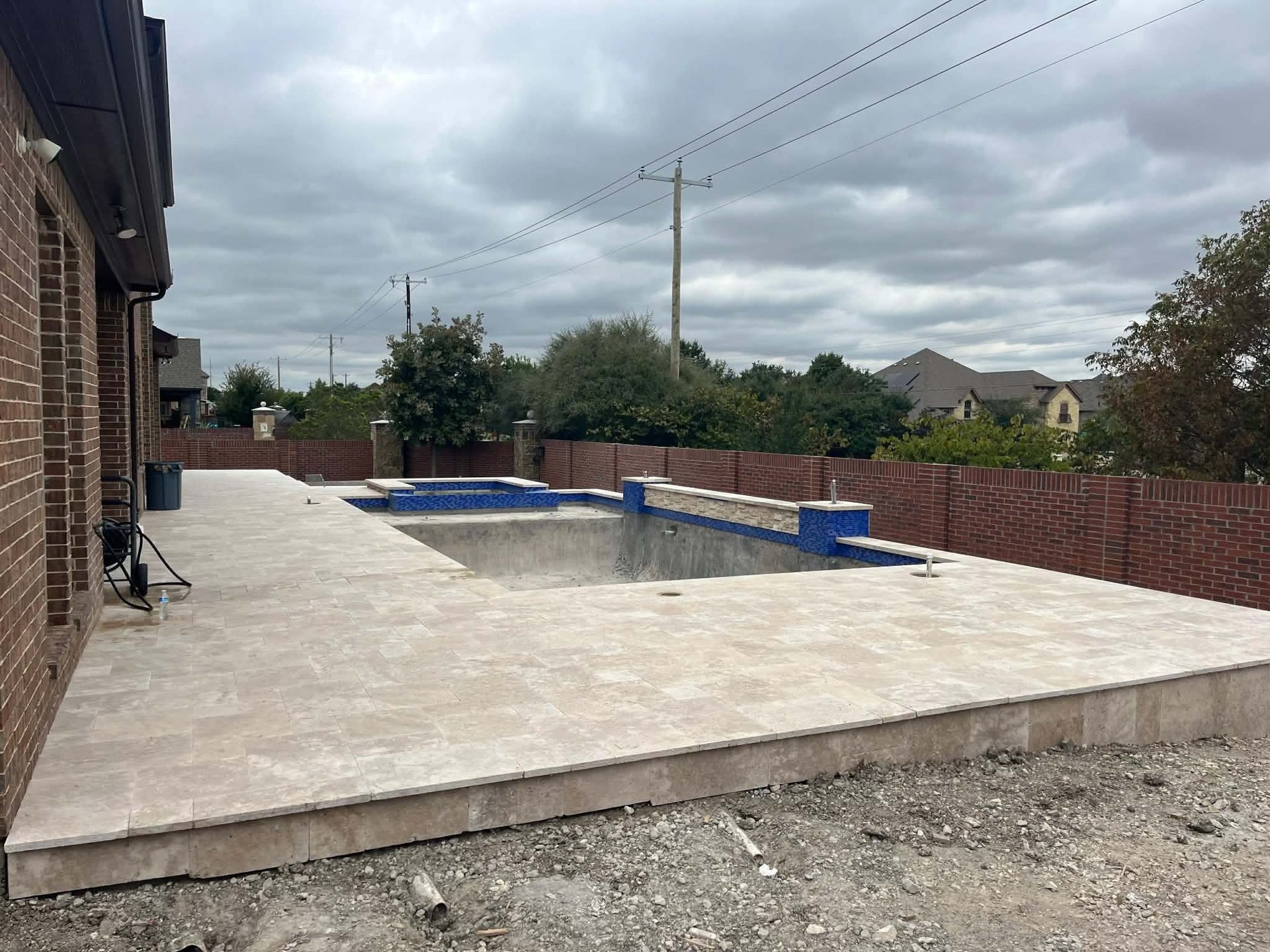 Residential backyard with concrete patio, swimming pool under construction with blue covers, red brick fence, and suburban homes visible in the background