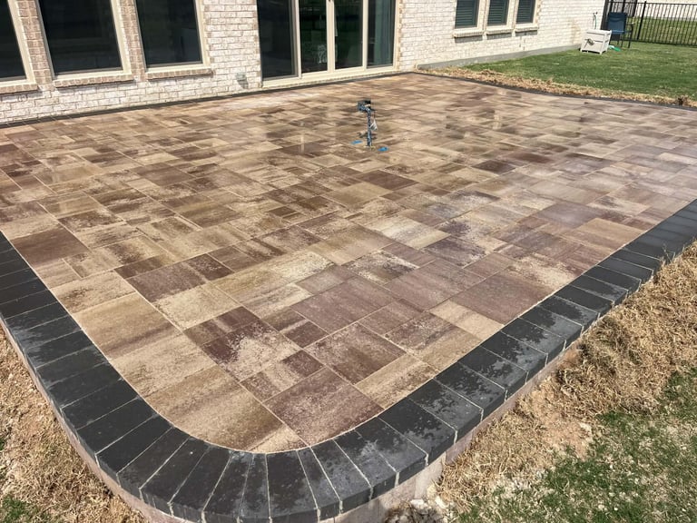Newly installed brown and tan brick patio with black border edging in backyard next to white brick house