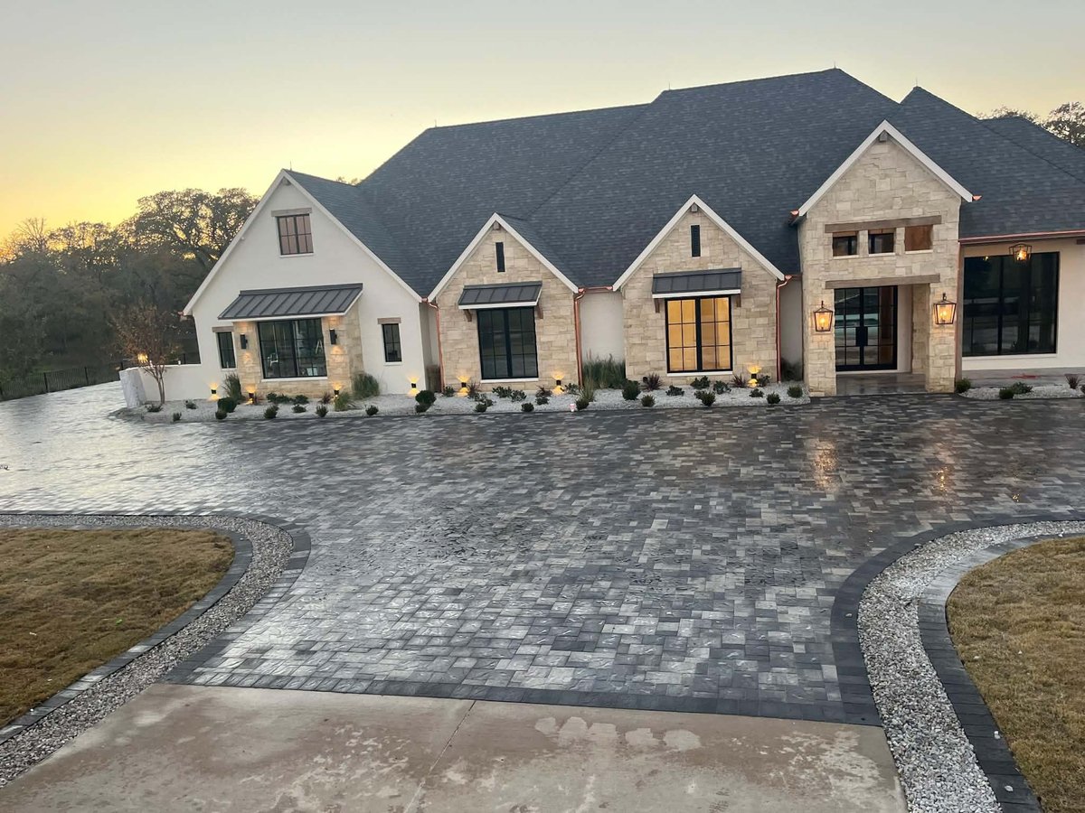 Modern two-story white brick home with dark roof and multi-car garage, featuring stone and paver driveway at dusk